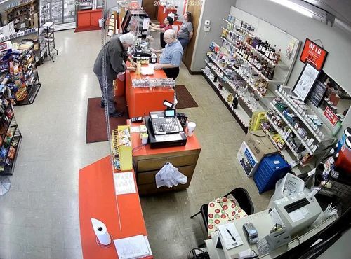 Convenience store checkout area with customers and clerks at orange counters, shelves lining the aisle.