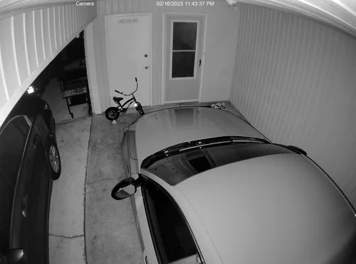 Surveillance view of a carport with two parked cars, a bicycle near the back wall, and a closed door.