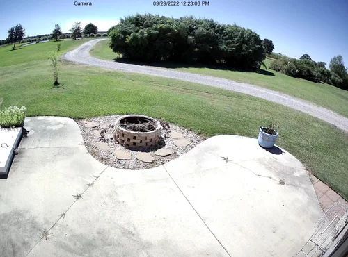 Sunny front porch overlooking a grassy lawn, winding driveway, and trees; fire pit in the foreground.