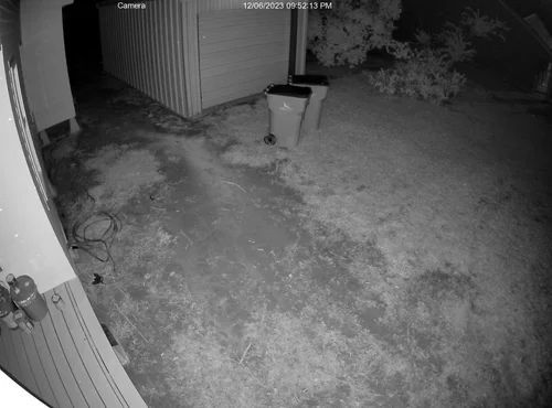 Night-vision view of a backyard beside a shed, with a trash bin and outdoor siding visible.