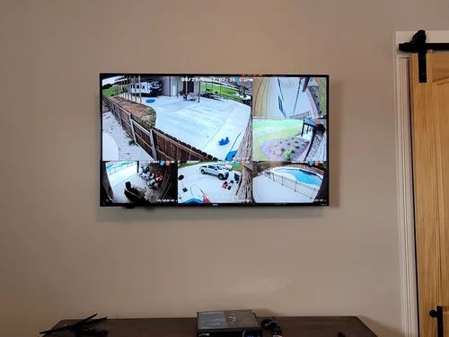 Wall-mounted TV showing a split-screen security camera view above a wooden table in a room