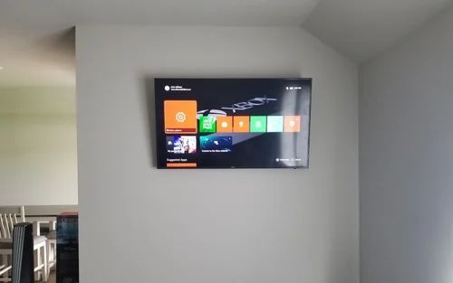 Wall-mounted TV displaying a colorful home screen on a gray wall in a room with tables and chairs.