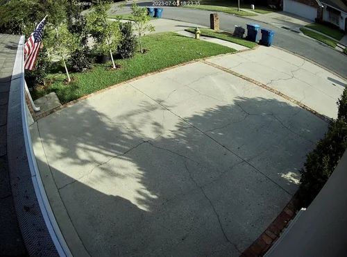 Driveway and front yard with lawn, trees, and an American flag beside a quiet street.
