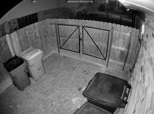 Night-vision view of a fenced backyard with a gate, trash bins, and a covered grill.