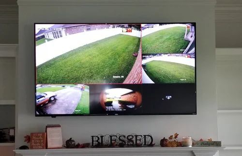 Wall-mounted TV showing four security camera views over a living room mantel with “BLESSED” decor