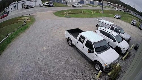 White pickup trucks parked on a gravel driveway beside a grassy yard and road.