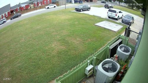 Residential courtyard with green lawn, parked cars, and trash bins beside a fence.