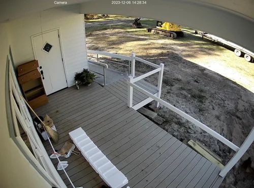 Front porch with white railing, chair, door, potted plant, and construction equipment in the yard beyond.