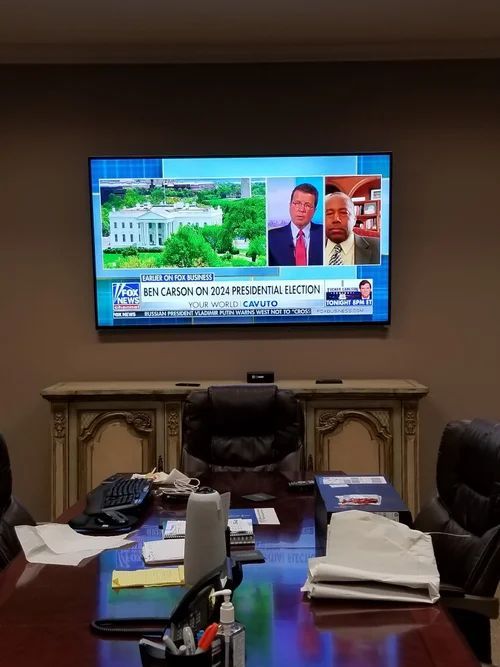 Conference room with a wall-mounted TV showing a news broadcast above a long meeting table.