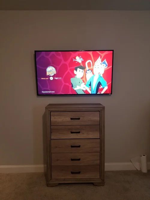 Wall-mounted TV showing a colorful cartoon above a wooden dresser in a bare room