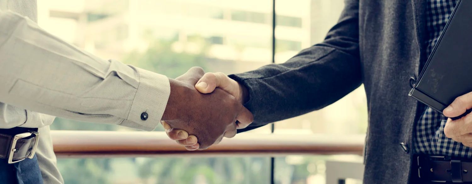 Two people shaking hands, one in a light jacket and one in a dark suit, in an office setting