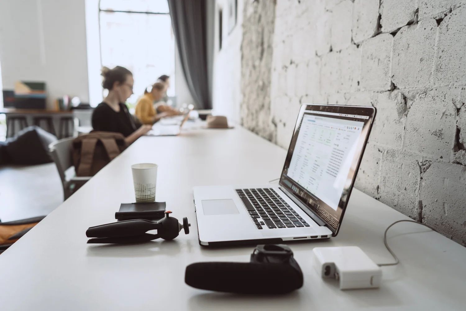 Laptop on a white desk in a bright office, with a phone, cup, and seated person in the background