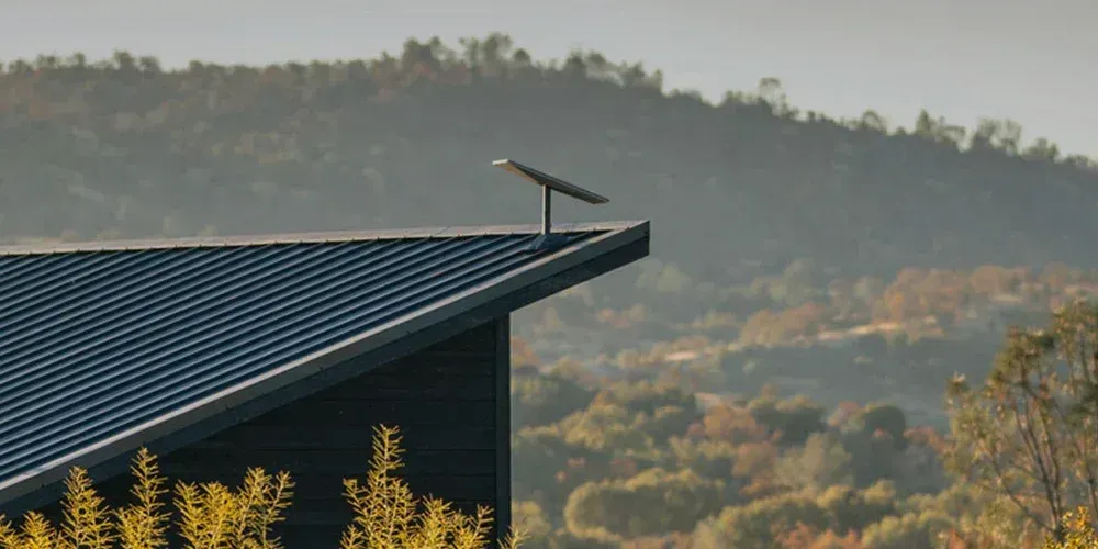 Slanted dark roof with a solar panel overlooking a hazy, tree-covered hillside at sunset