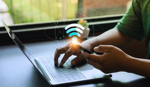 Person using a laptop and smartphone beside a glowing Wi‑Fi icon on a desk near a window