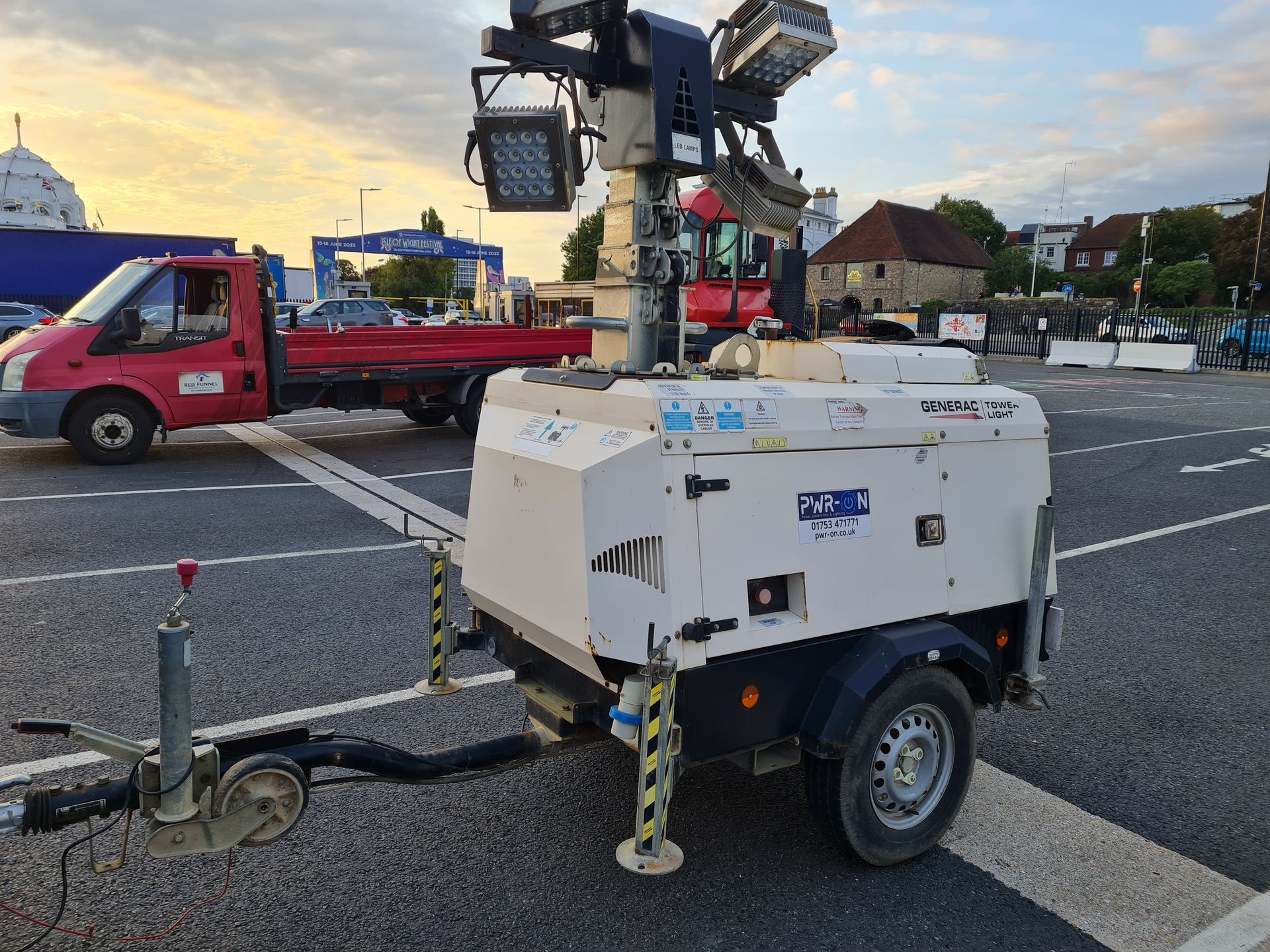 White portable light tower on a trailer in a parking lot, with a red tow truck nearby at dusk