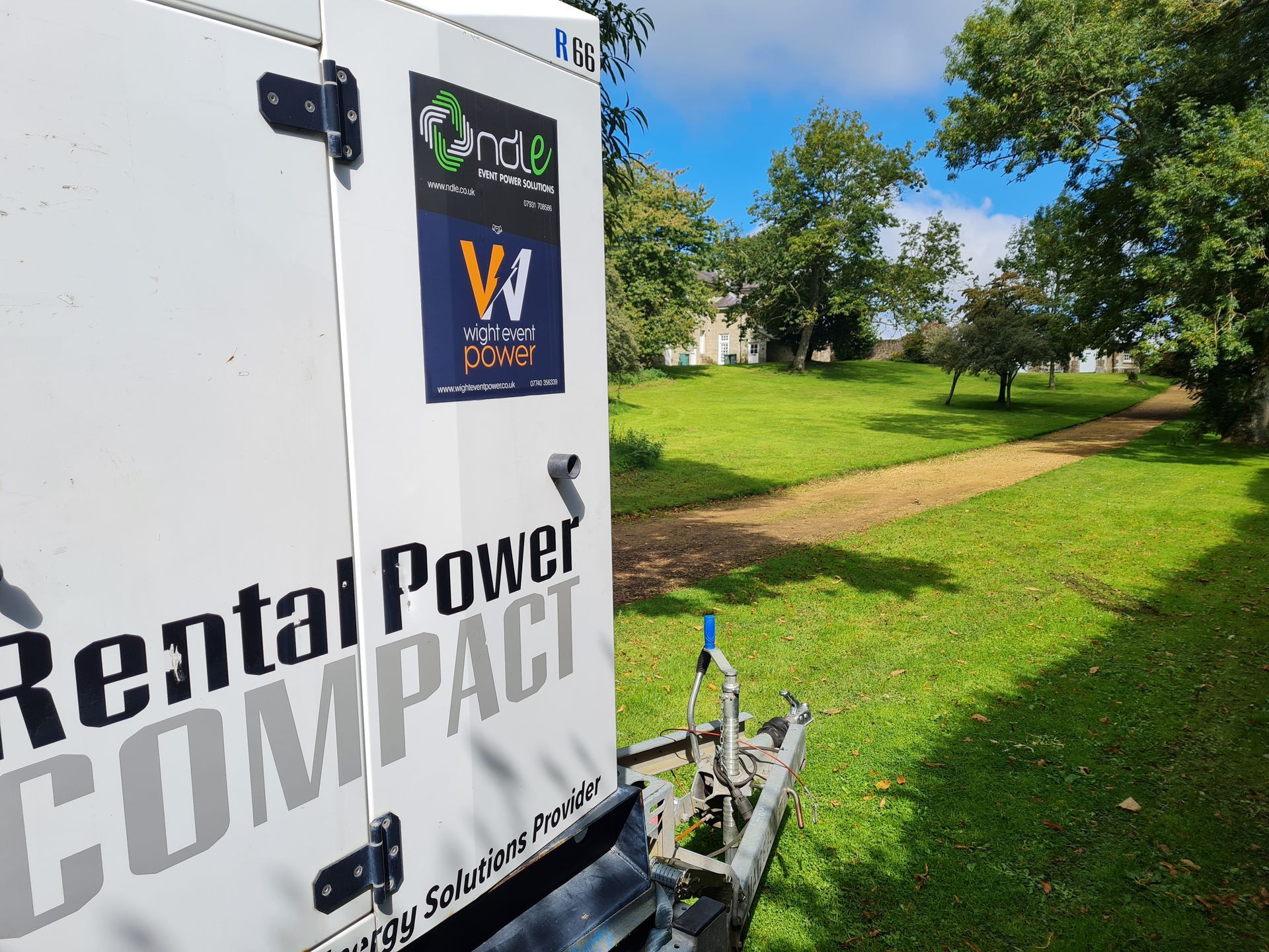White Rental Power Compact trailer parked on grass beside a sunny park with trees and a signboard