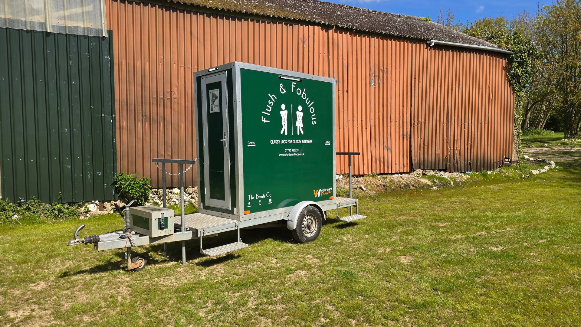 Green portable toilet trailer on grass beside a wooden fence under a sunny sky.