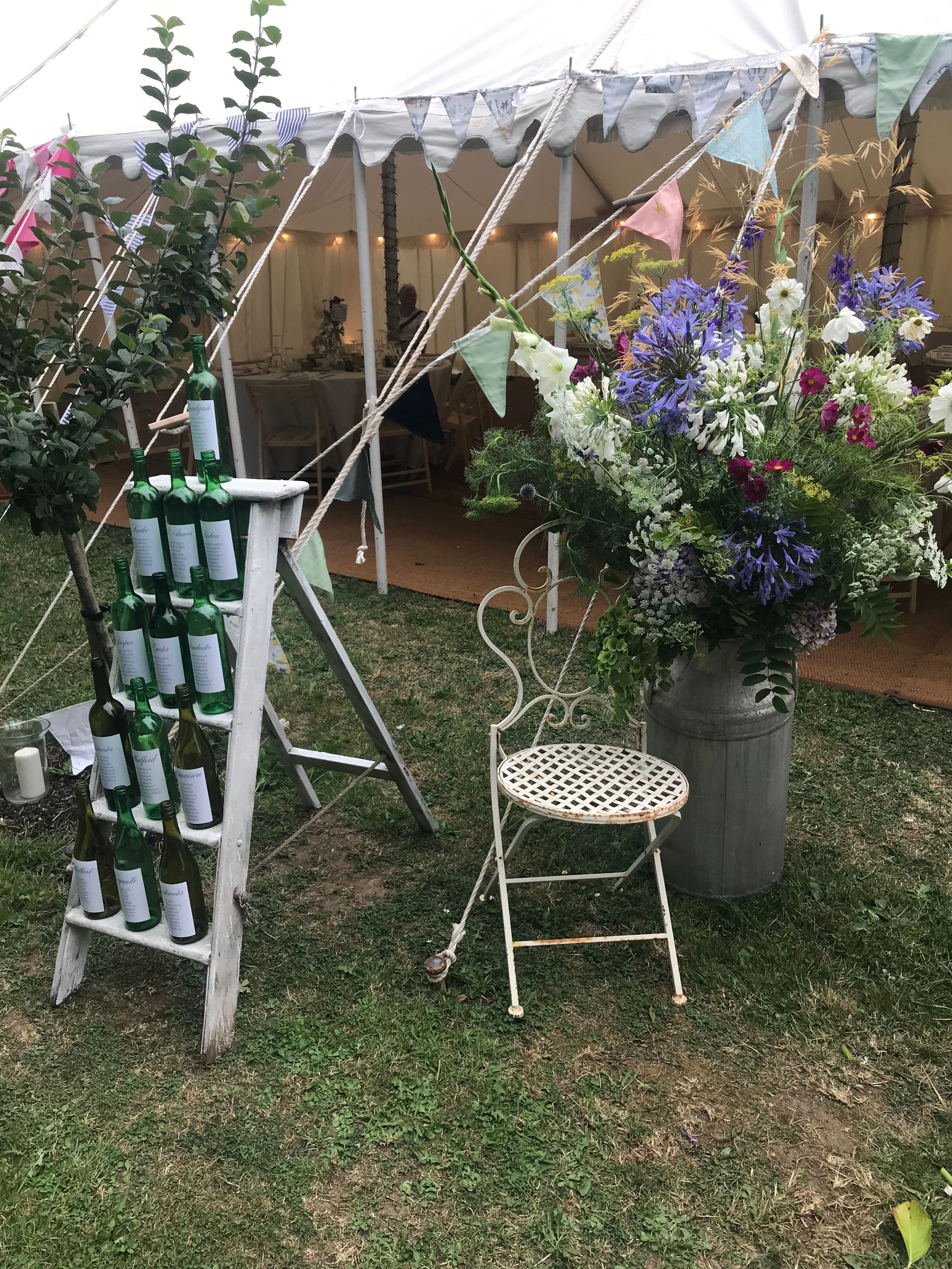 Outdoor floral display with potted purple and white flowers beside a small white chair and trellis ladder under a tent