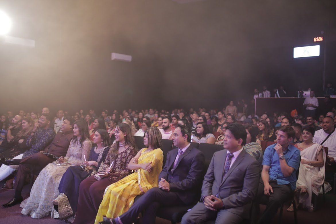 Audience seated in a dim auditorium, watching a performance. Stage is obscured by light and smoke.