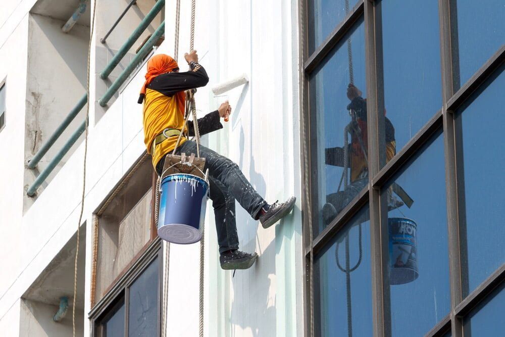A Man is Painting a Building With a Roller While Hanging From a Rope — NHM Painting & Projects in Ciccone, NT