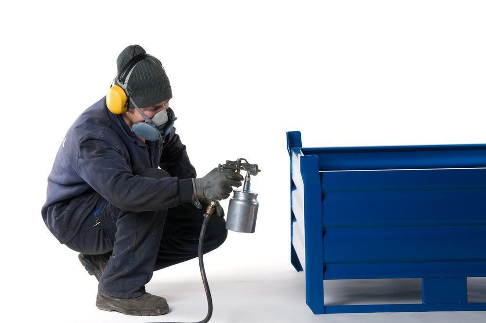 A Man Wearing a Mask and Ear Muffs is Spray Painting a Blue Box — NHM Painting & Projects in Ciccone, NT