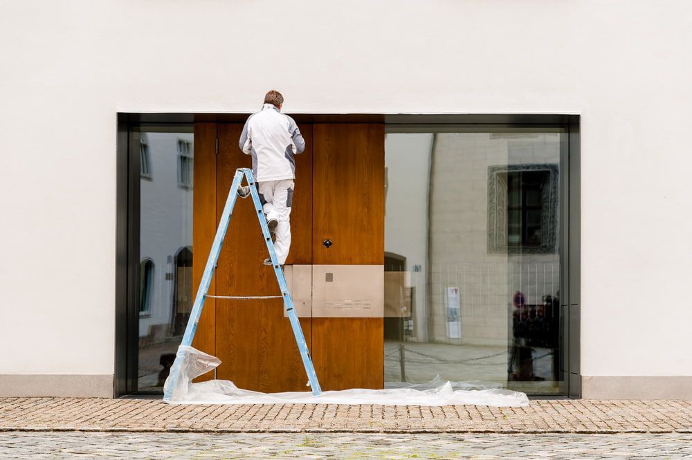 A Man is Standing on a Ladder Painting a Door — NHM Painting & Projects in Ciccone, NT