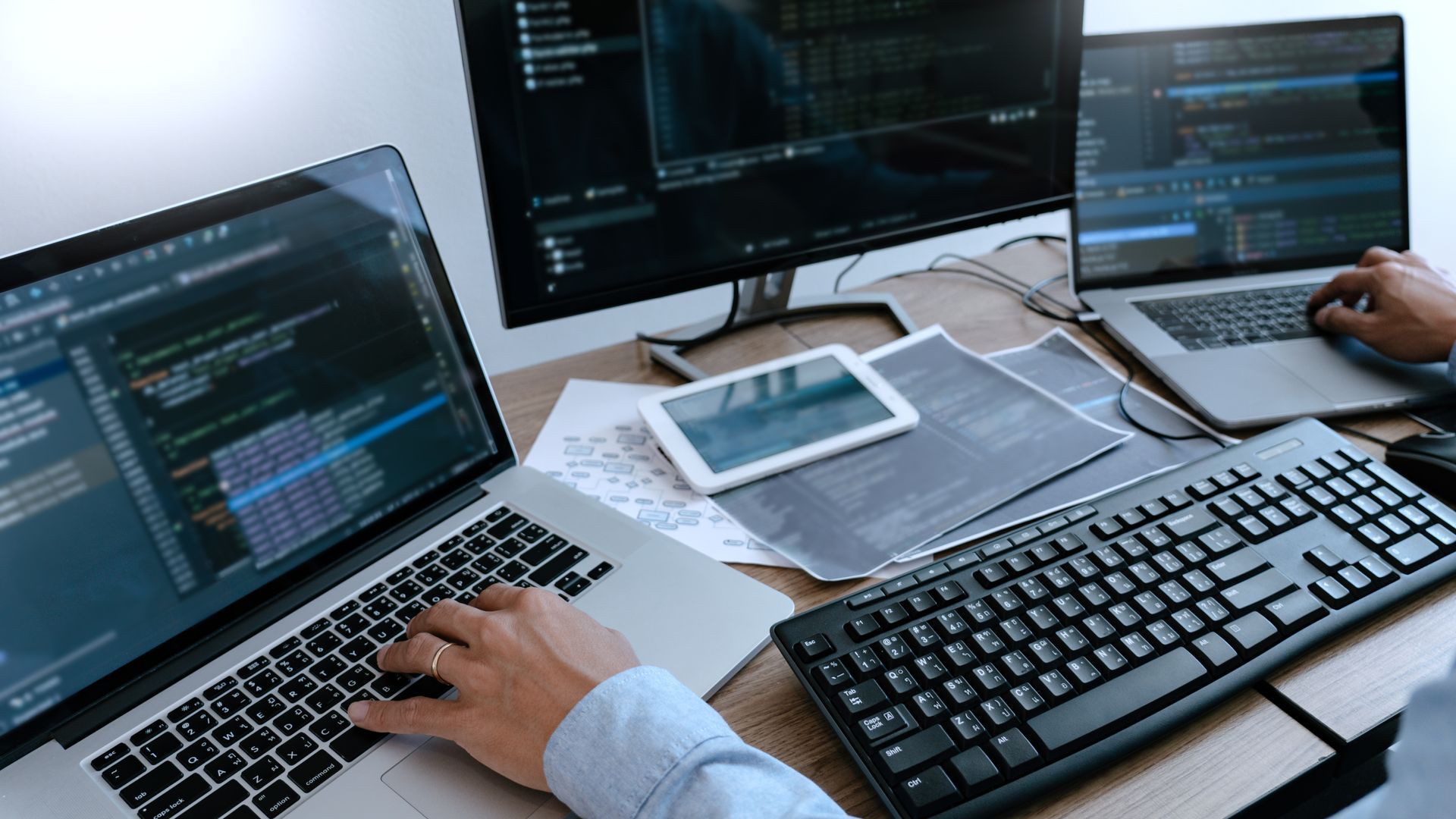 Hands typing on laptops at a desk with code on multiple screens and a tablet in the center