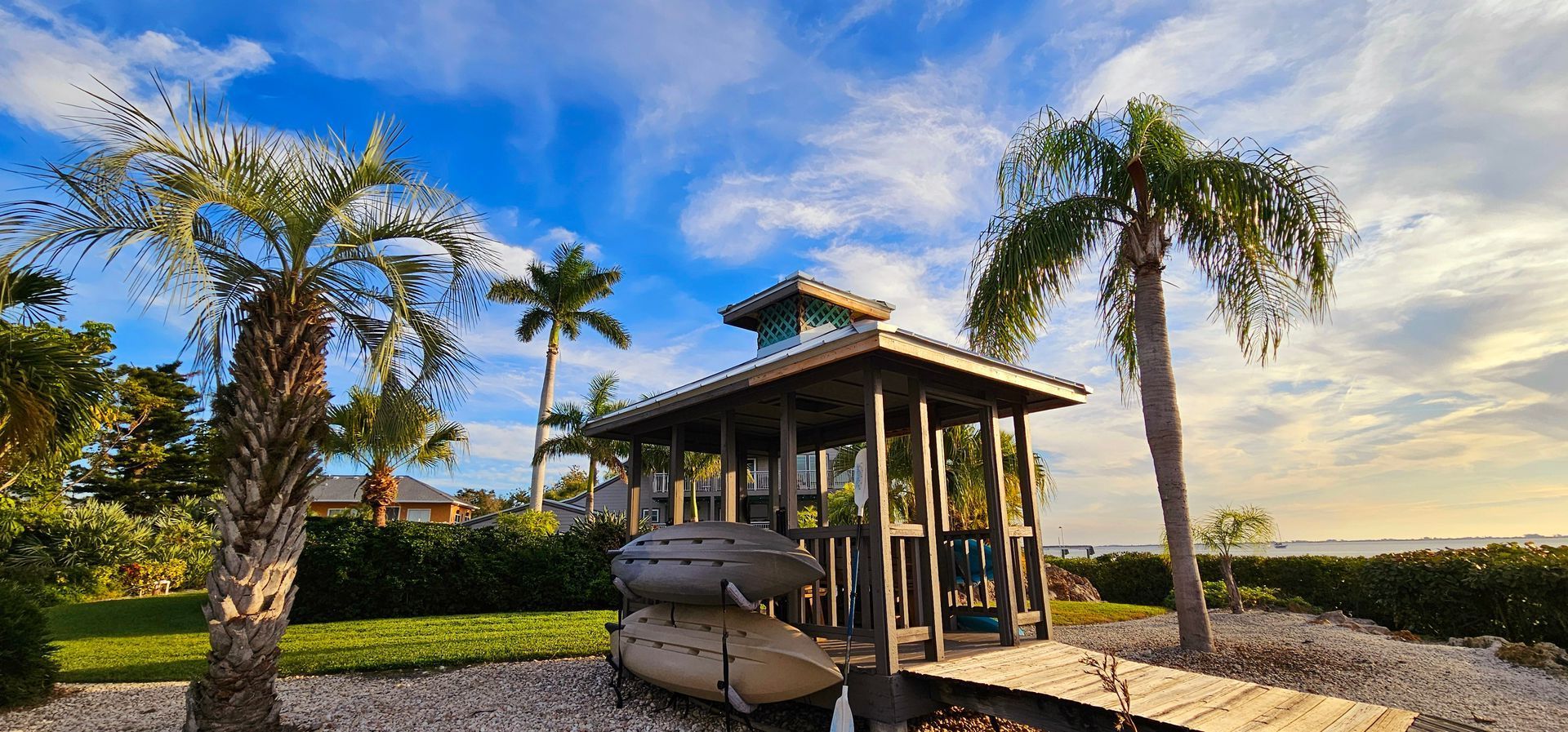 Tropical beachside gazebo with palm trees, blue sky, and surfboards on sand at sunset