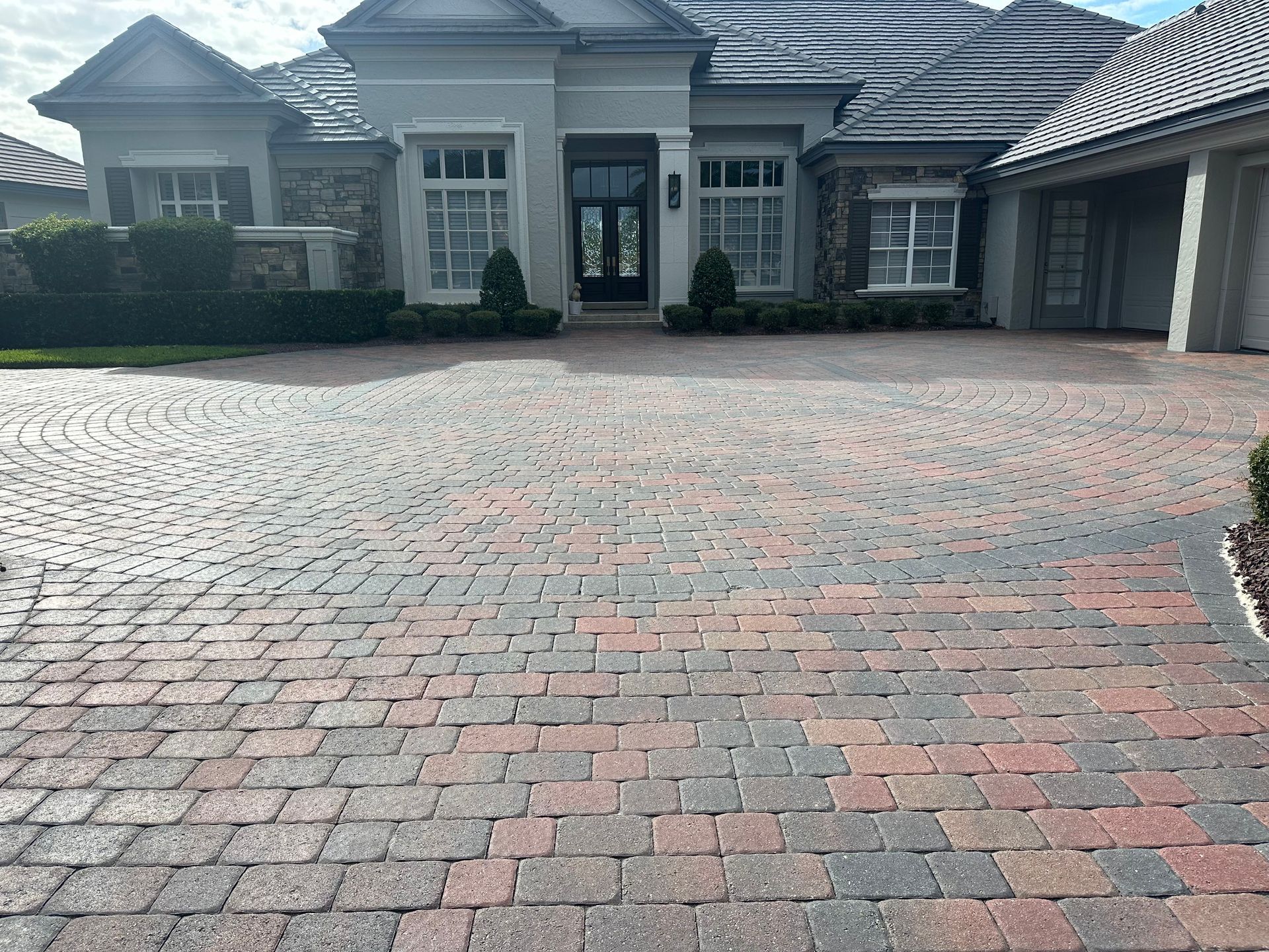 Brick driveway leading to a light-colored house with arched windows and a dark door.