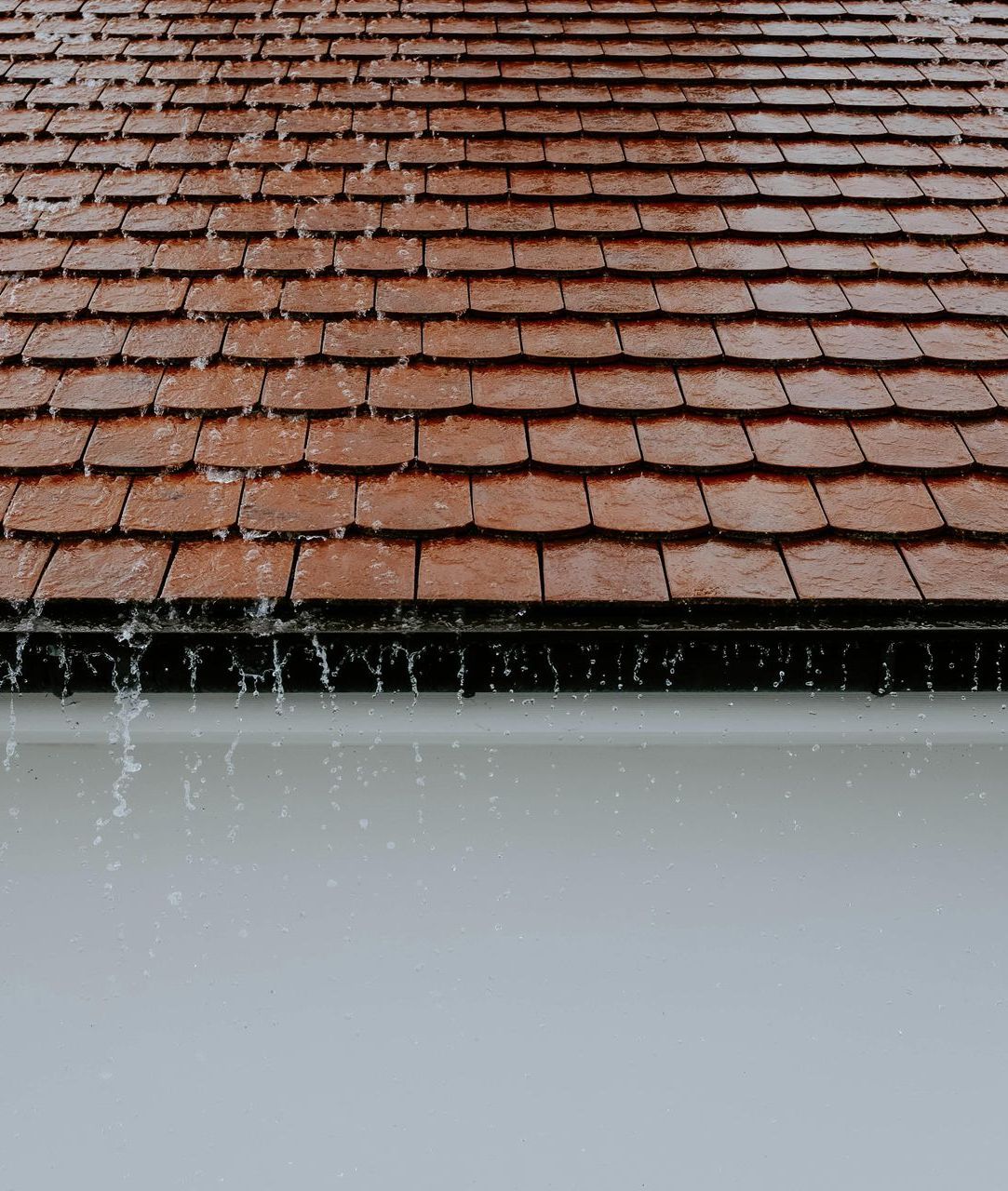 Rainwater dripping from a red tiled roof over a white wall.