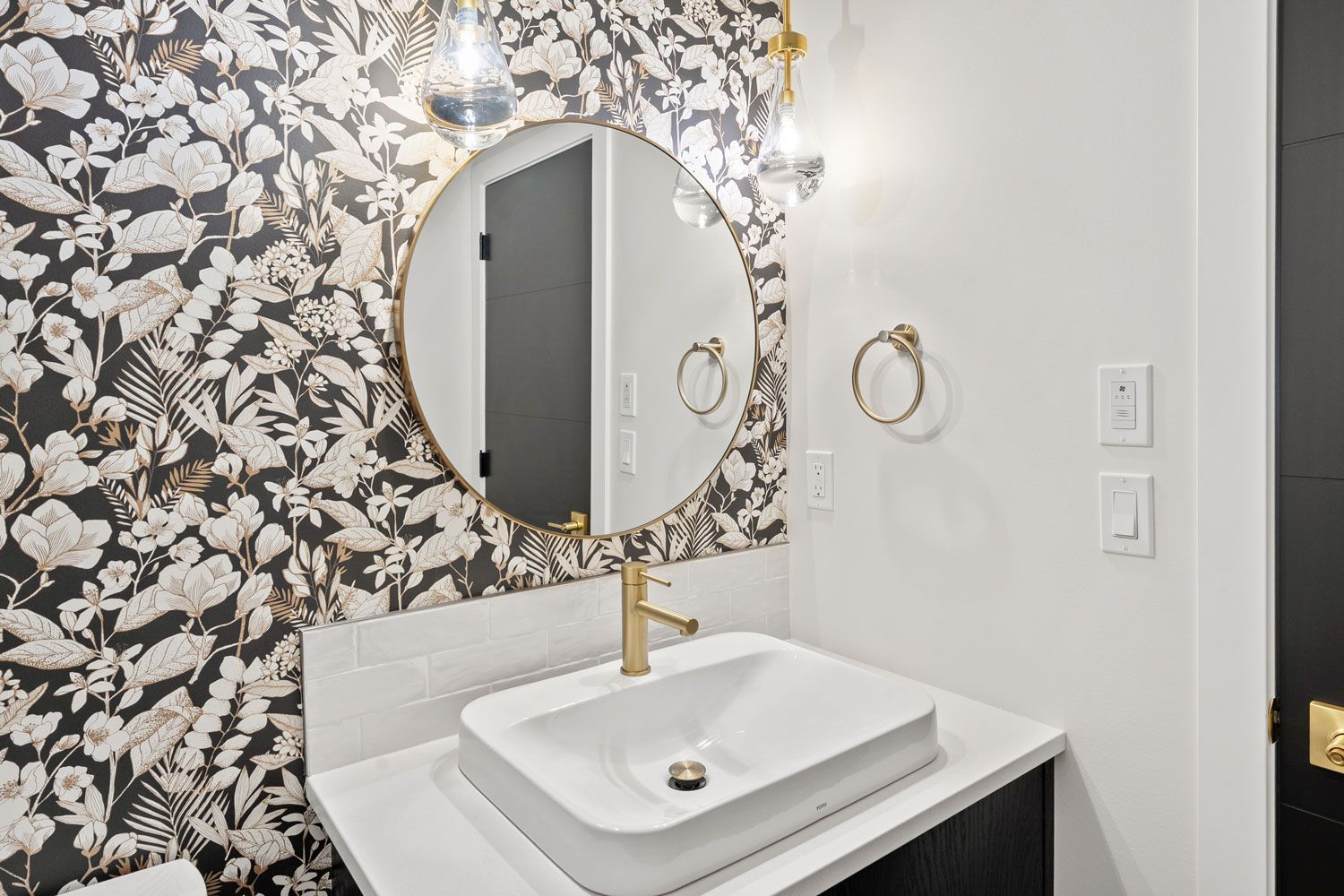 Modern bathroom vanity with round mirror, floral wallpaper, and white sink with gold accents