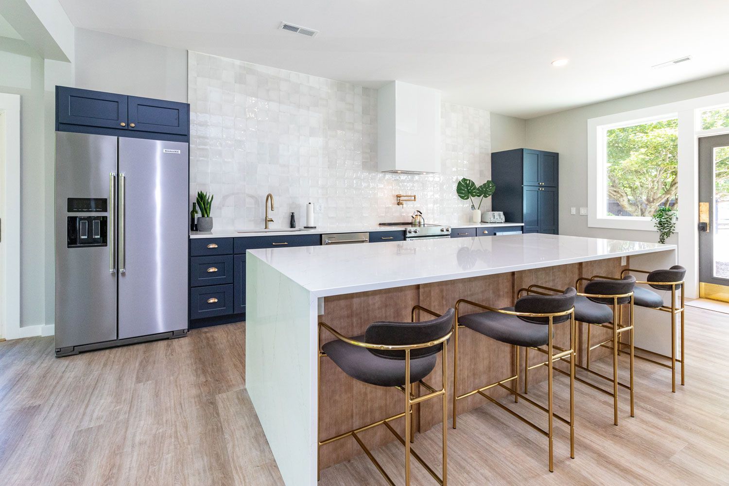 Modern kitchen with white island, gold stools, stainless fridge, navy cabinets, and sliding glass doors
