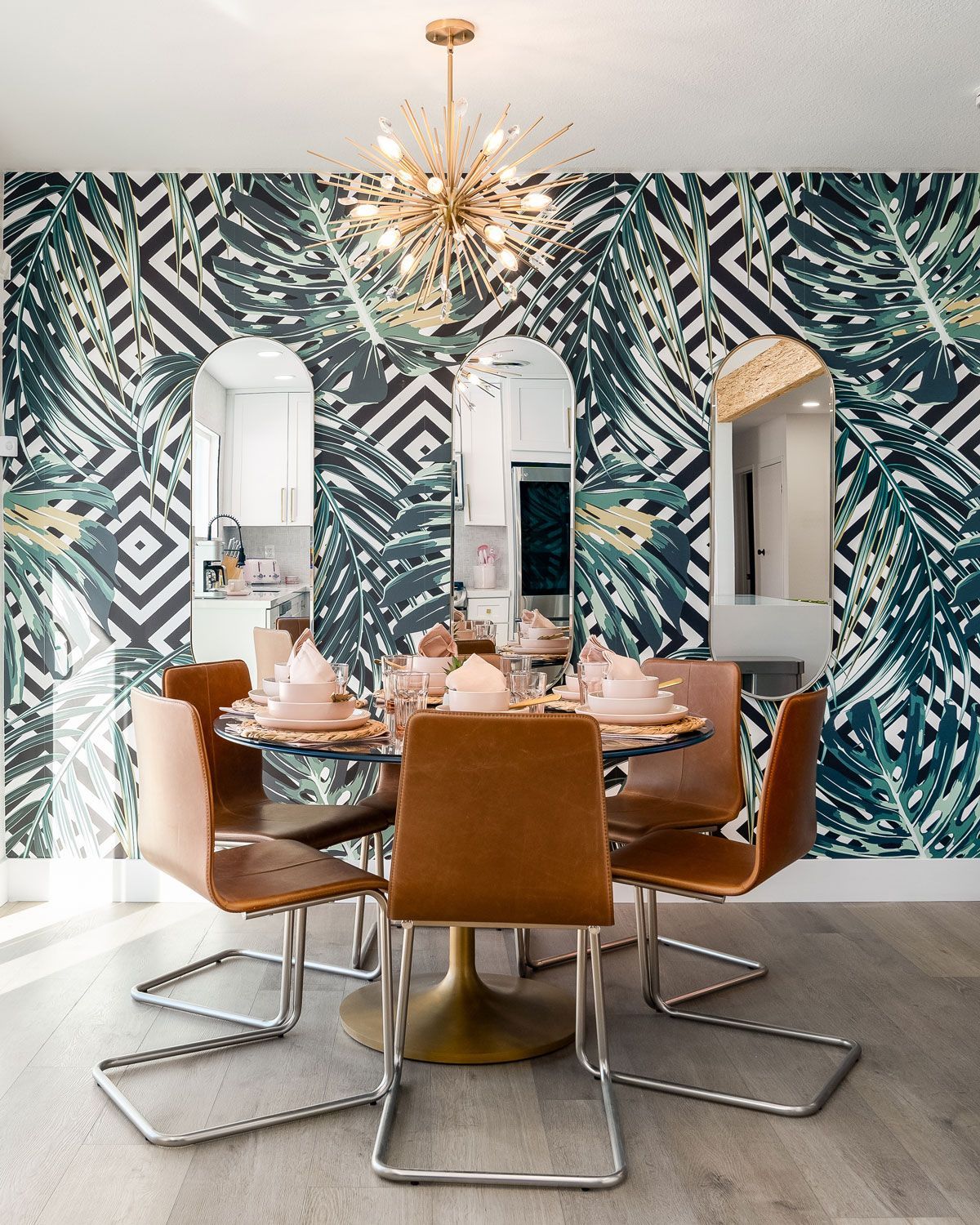 Modern dining room with geometric wallpaper, gold chandelier, and glass table with tan chairs.