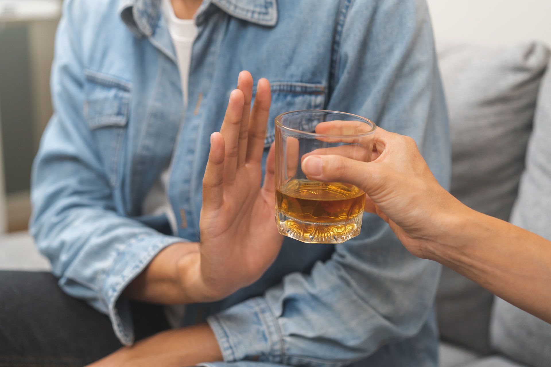 A person in a denim shirt holds up a hand to refuse a glass of amber-colored liquid offered by another person.