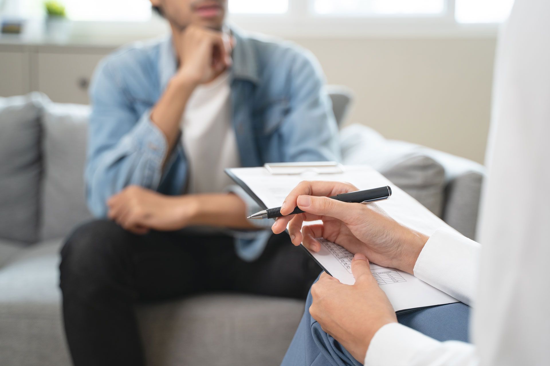 A person holds a pen and clipboard, recording notes while another person sits in the background on a sofa.