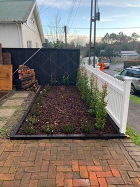 Freshly planted narrow garden bed beside a brick driveway and white fence, with a black privacy fence at the end.