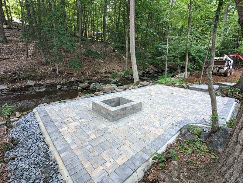 Paver patio with fire pit next to a stream in a wooded area.