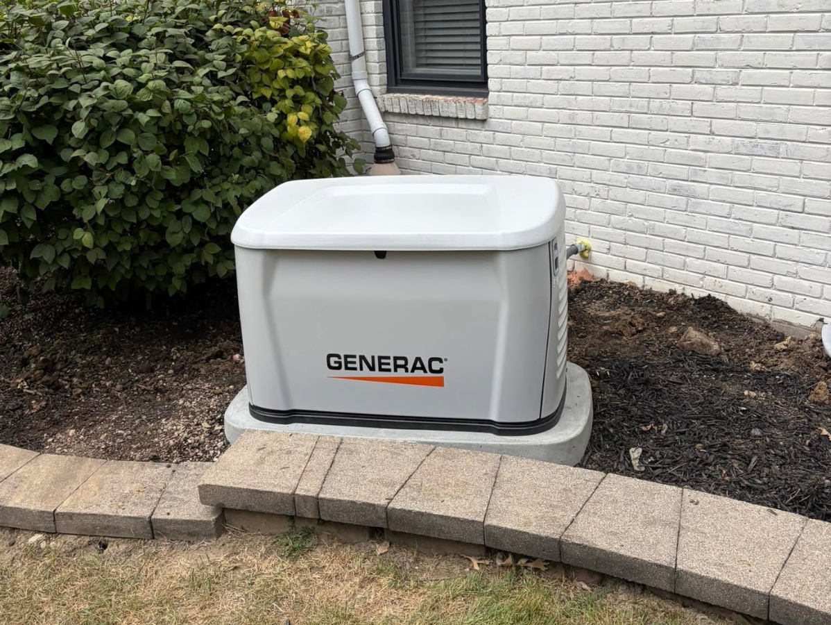 Generator near a white house, on a concrete pad. Electrical box on the wall. Brown, green surroundings.