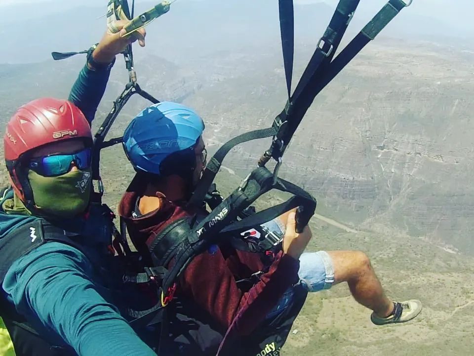 Dos personas volando en parapente sobre un paisaje montañoso; una con un casco rojo y la otra con un casco azul.