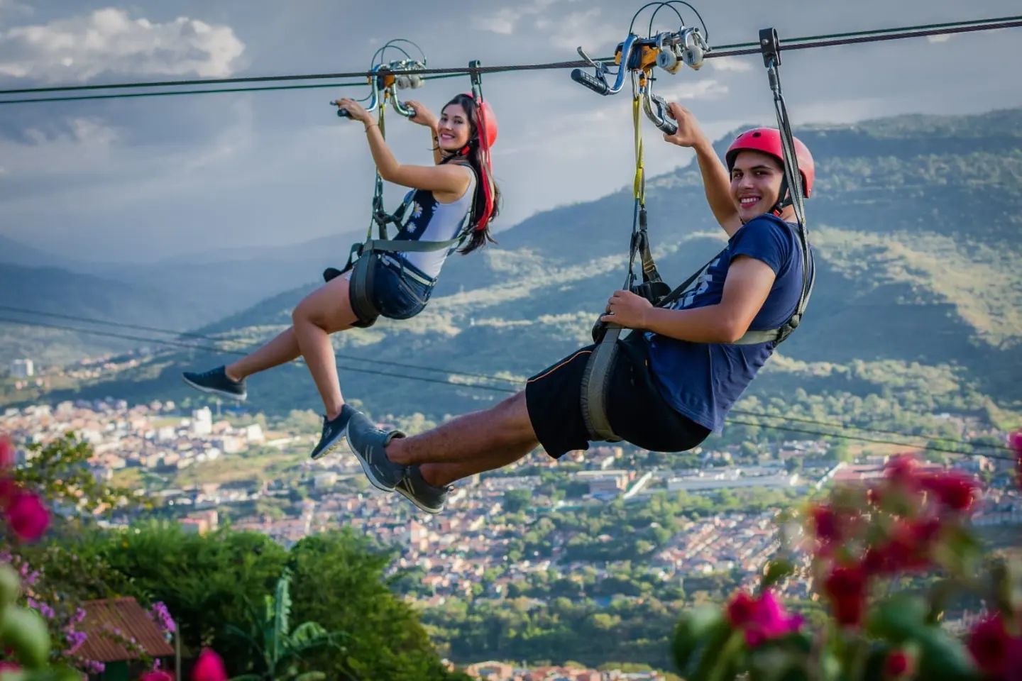 Dos personas haciendo tirolesa con un paisaje urbano de fondo; llevan cascos y sonríen.