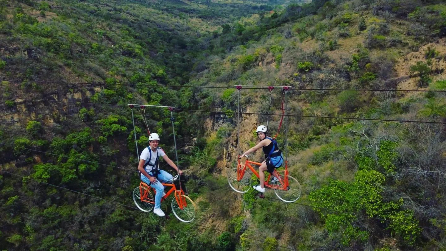 Dos personas viajan en bicicletas naranjas suspendidas sobre un cañón, rodeadas de árboles verdes y follaje.