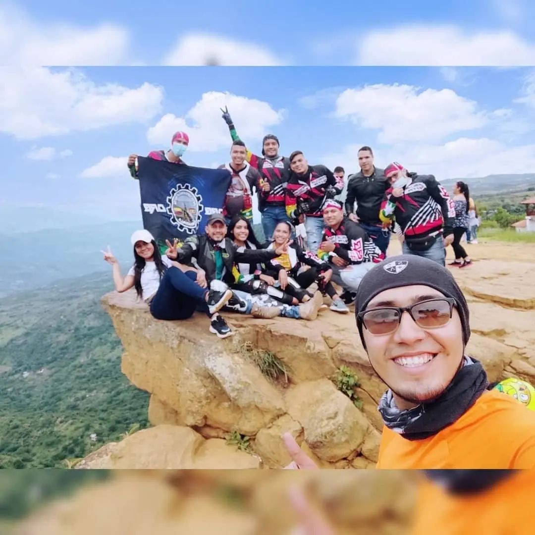 Grupo de personas en un acantilado, con una bandera, sonriendo. Montaña y cielo al fondo.