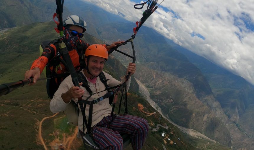 Dos personas en parapente sobre un valle montañoso en un día soleado. Una con casco naranja y la otra con casco blanco.