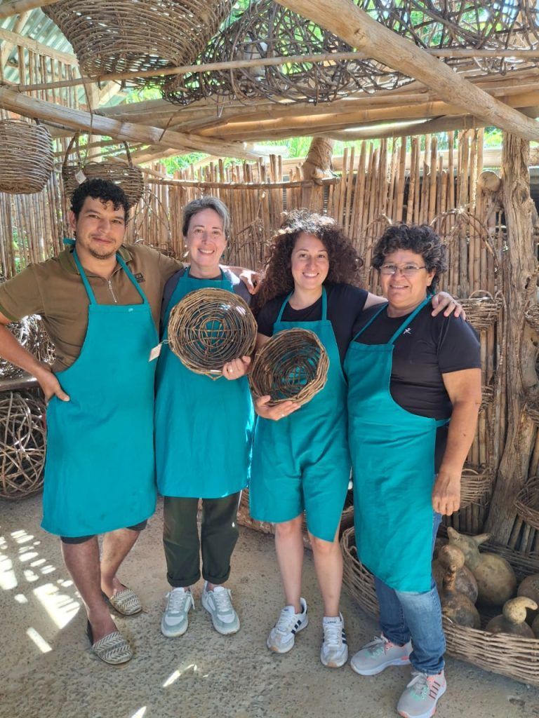 Cuatro personas con delantales verde azulado sostienen cestas tejidas y sonríen en un taller al aire libre.