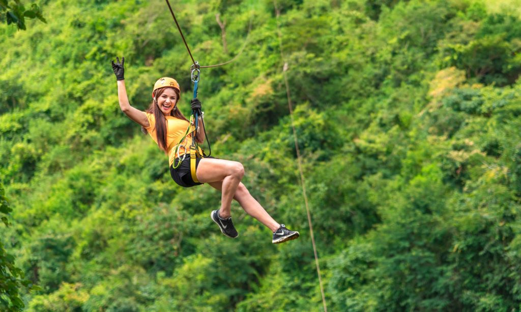 Mujer haciendo tirolesa a través de un exuberante bosque verde, sonriendo y saludando.