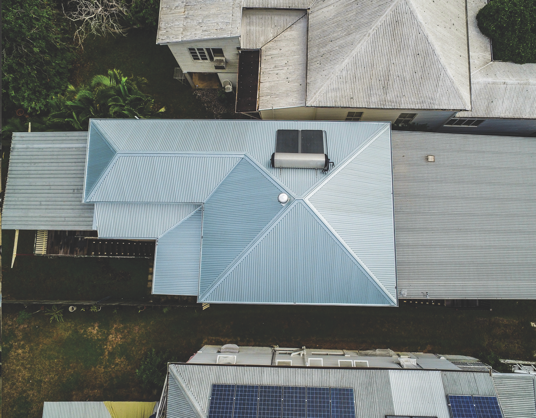 Aerial view of a house with a light blue metal roof surrounded by neighboring roofs and trees