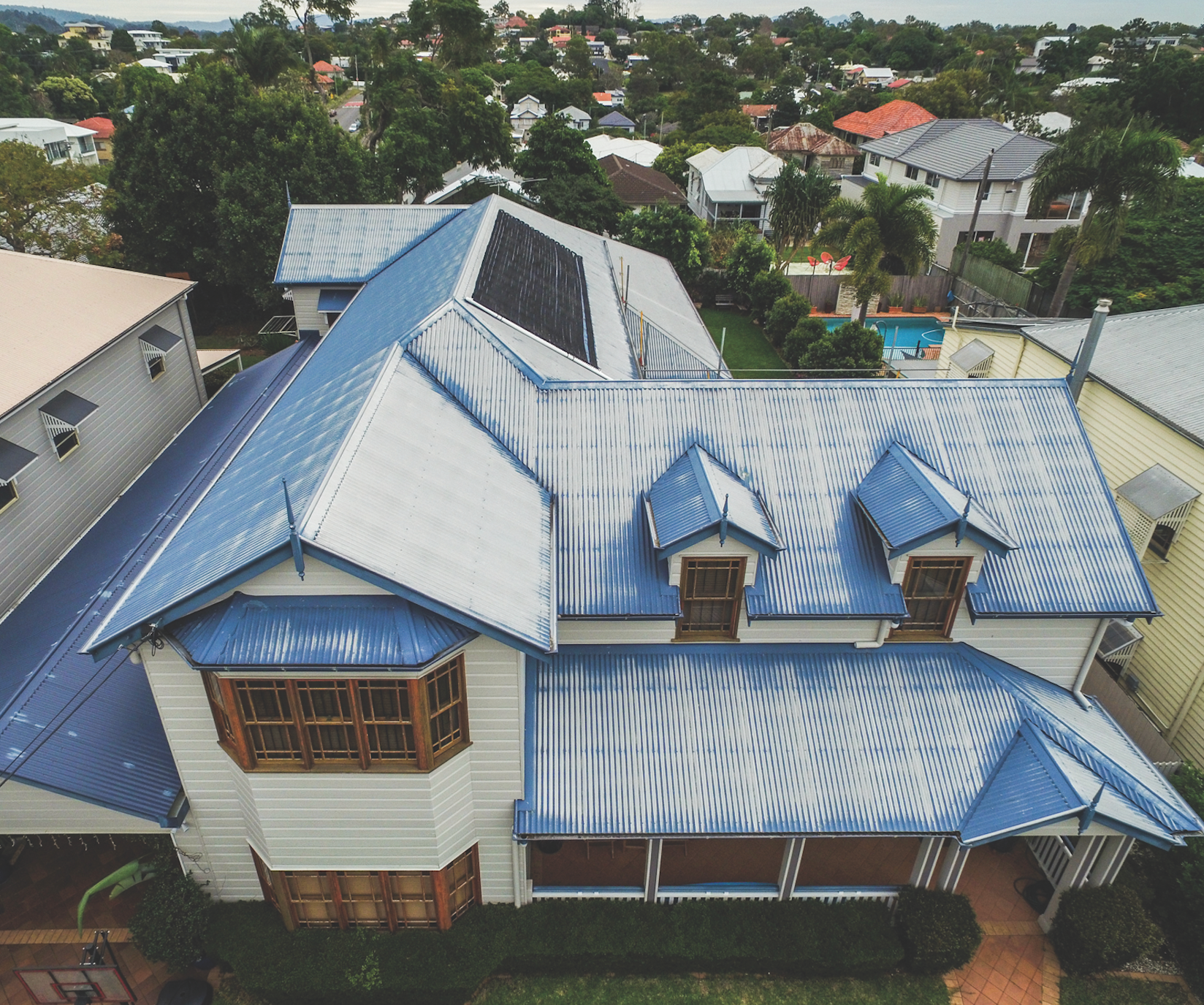 Aerial view of a large house with blue metal roof in a leafy suburban neighborhood