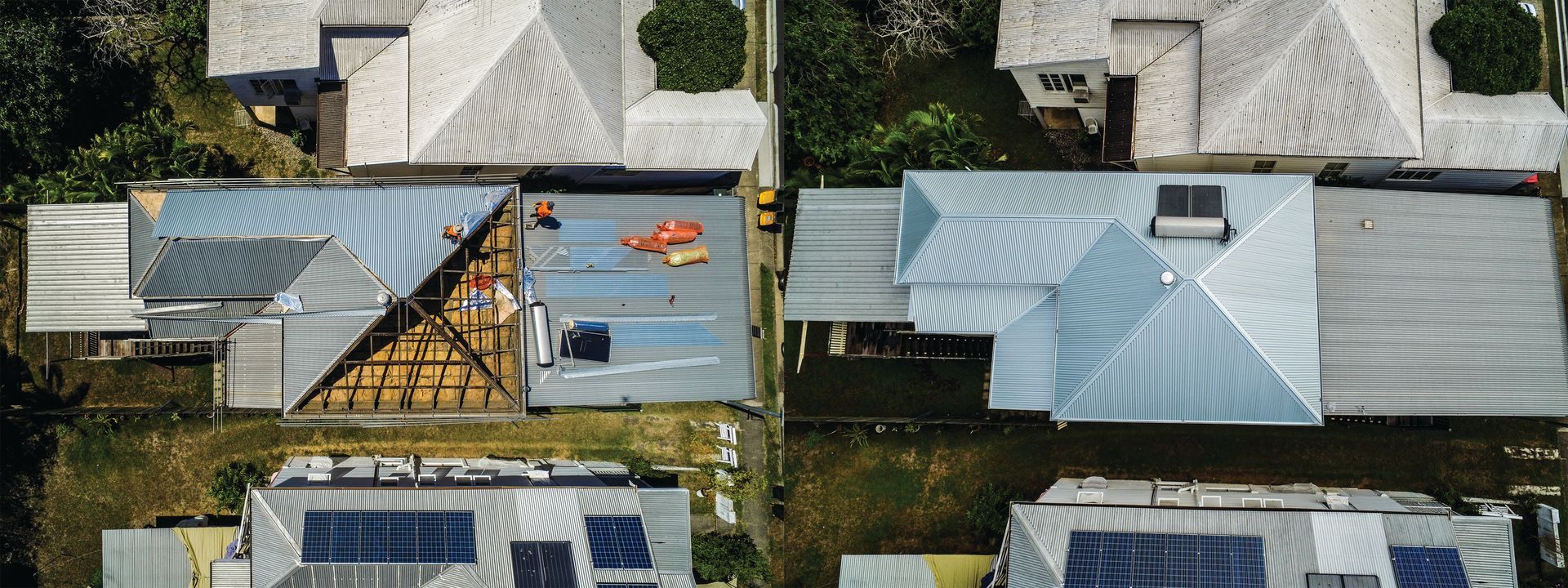 Aerial view of two homes with blue roofs, solar panels, and people on a rooftop deck by a pool.