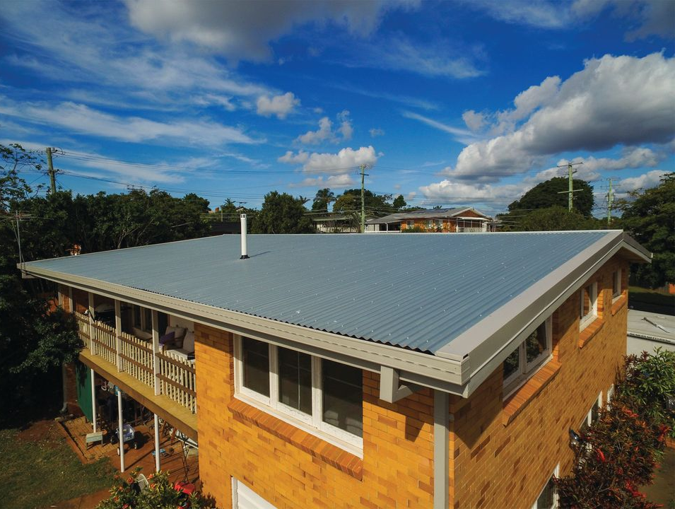 House with gray metal roof under a bright blue sky and clouds, seen from above with trees nearby
