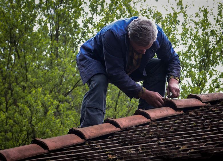 Worker in a blue jacket repairing red roof tiles beside green trees.